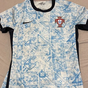 Portugal Nike fotbollströja blåvit - Säljer en Portugal fotbollströja från Nike med Dri-FIT ADV-teknologi. Tröjan är vit med ljusblå geometriskt mönster, har svarta detaljer vid krage och ärmslut samt Portugal-emblem på bröstet. Kortärmad och tillverkad i lätt, ventilerande material.