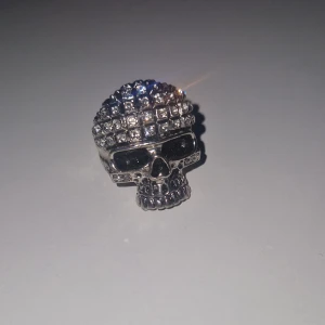 Dödskallering med stenar - Cool ring i form av en dödskalle, silverfärgad med glittrande stenar på huvudet och svarta detaljer vid ögonen. Perfekt statement piece för dig som gillar edgy stil och vill sticka ut. Ringen har en robust design och är riktigt iögonfallande.