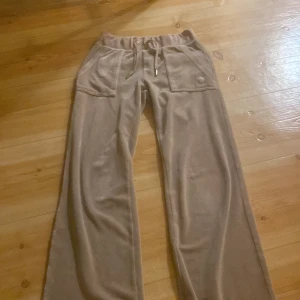 Beiga velourbyxor från Juicy Couture - Supermjuka beiga velourbyxor från Juicy Couture med bred resår i midjan och snörning med metalländar. Byxorna har stora fickor fram och bak med knappar och broderad JC-logga på ena fickan. Perfekta för en chill och trendig look. Köpt ifrån bubbleroom! Storlek xxs!