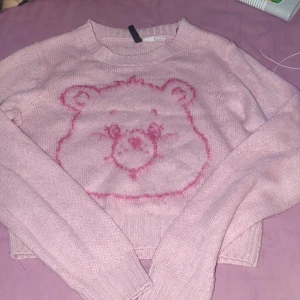 Rosa stickad tröja med björnansikte - Supergullig ljusrosa stickad tröja från Care of Wear i storlek M. Tröjan har ett stort björnansikte i rosa framtill, ribbade muddar och lång ärm. Perfekt för dig som gillar söta detaljer och vill ha en mysig vibe.