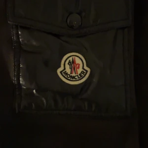 Svart pufferjacka från Moncler - Svart cardigan från Moncler med huva och klassisk logga på bröstfickan. Jackan har quiltade ärmar i glansigt material, dragkedjor på sidorna och en snygg snörning till huvan. Perfekt för dig som vill ha en stilren och exklusiv look.