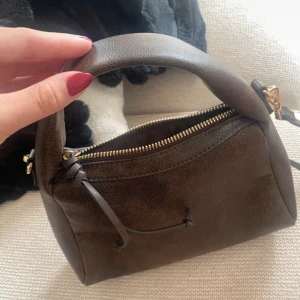 Brun handväska i skinn med dragkedja - Snygg brun handväska i skinn med både handtag och axelrem. Väskan har en stilren design med guldfärgade detaljer och dragkedja upptill. Perfekt storlek för det viktigaste och passar till många olika outfits. Helt ny!
