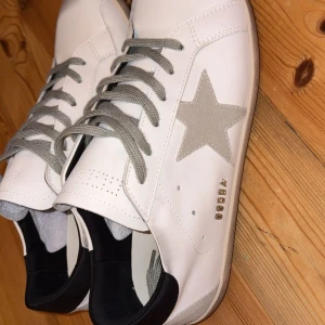 Golden Goose  - Golden Goose Superstar sneakers i vitt skinn med grå mockadetaljer och grå snörning. Skorna har en ikonisk stjärna i mocka på sidan, svart hälparti och slitna detaljer på sulan för en cool look. Perfekta för dig som gillar streetstyle och unika detaljer.
