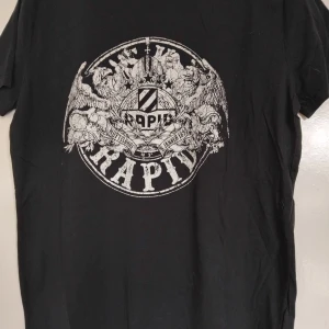 Svart Rapid Wien t-shirt med tryck - Cool svart t-shirt från Rapid Wien med stort vitt tryck på bröstet som visar klubbens emblem och detaljerade grafiska inslag. Rund hals och korta ärmar, perfekt för dig som vill visa ditt stöd för laget. Materialet är mjukt och bekvämt bomull.