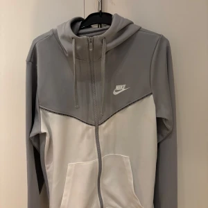 Grå och vit Nike hoodie med dragkedja - Nike hoodie i grått och vitt med hel dragkedja framtill och huva med snörning. Tröjan har fickor på magen och Nike-logga på bröstet. Perfekt för en sportig och avslappnad stil.