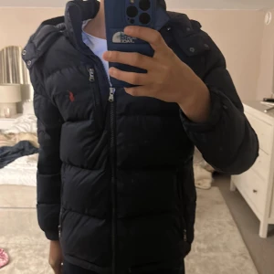 Blå dunjacka från Polo Ralph Lauren - Säljer en blå dunjacka från Polo Ralph Lauren med röd logga på bröstet. Jackan har huva, två sidofickor med dragkedja och en hög krage. Materialet är syntet och dun, perfekt för kalla dagar. Stängs med dragkedja framtill och har en klassisk, puffad look. Storlek L i barn (14 - 16). Ny pris 3000 kr.
