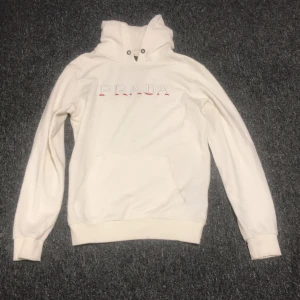 Vit hoodie från Prada med broderi - Exklusiv vit hoodie från Prada i storlek L. Tröjan har broderad Prada-logga framtill i vitt och rött, huva med snörning och klassisk känguruficka. Mjuk bomullskänsla och ribbade muddar vid ärmslut och nederkant. Perfekt för dig som gillar stilrena designer.