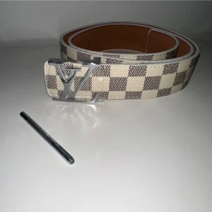 Louis Vuitton rutig beige bälte - Snyggt bälte från Louis Vuitton med klassiskt rutigt mönster i beige och brunt. Bältet har en stor silvrig LV-logga som spänne och är gjort i skinn. Perfekt accessoar för att lyfta din outfit.
