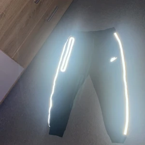 Svarta Nike mjukisbyxor med reflex - Svarta mjukisbyxor från Nike med feta reflexdetaljer längs benen och en reflekterande Nike-logga på låret. Byxorna har resår i midjan och muddar vid bensluten. Perfekta för kvällspromenader eller chill dagar.