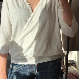 Vit omlottopp med trekvartsärm - Snygg vit omlottopp med v-ringning och trekvartsärmar. Toppen har en avslappnad passform och är tillverkad i mjukt bomullsmaterial. Perfekt att styla med jeans för en enkel men trendig look.