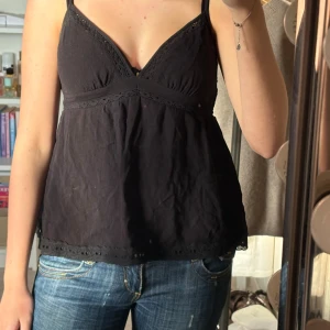 Svart linnetopp med spetsdetaljer - Svart linnetopp med smala axelband och v-ringning. Toppen har fina spetsdetaljer längs ringningen, under bysten och vid fållen. Lätt och luftig modell som passar perfekt till jeans eller kjol. Materialet känns mjukt och bekvämt mot huden.