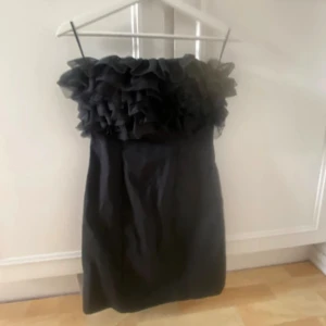 Svart kort klänning med volang - Supersnygg svart kort klänning med axelbandslös design och flera lager volanger upptill. Klänningen har en rak passform och är perfekt för dig som vill sticka ut. Materialet känns lätt och har en elegant struktur.