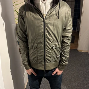 G star jacka - Snygg olivgrön bomberjacka från G-Star RAW med huva och dragkedja framtill. Jackan har två fickor med knappar, ribbade muddar och en sportig look. Perfekt för dig som gillar streetwear och vill ha en jacka som sticker ut. Jackan har ett hål i luvan