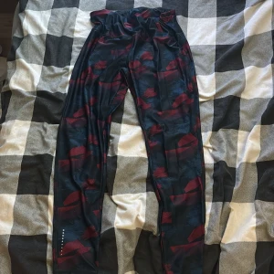 Mönstrade träningsbyxor från Crivit - Säljer ett par svarta och mörkblå träningsbyxor från Crivit med röda och blå mönsterdetaljer. Byxorna är tillverkade av återvunnet syntetmaterial och har en elastisk midja för skön passform. Perfekta för gymmet eller löprundan.
