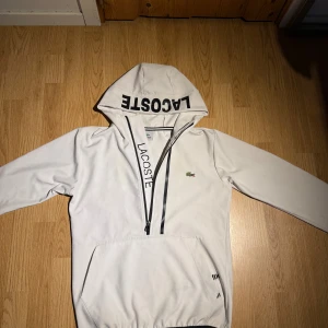 Vit hoodie från Lacoste med dragkedja - Säljer en stilren vit hoodie från Lacoste med svart logga på huvan och dragkedja framtill. Klassisk krokodil på bröstet, stor ficka framtill och långa ärmar. Perfekt för dig som gillar sportig och clean stil.