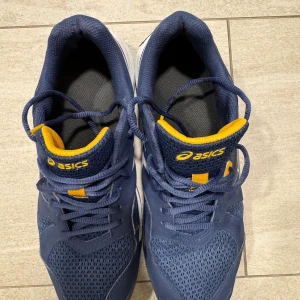 Blå Asics Gel-Padel Pro 5 sneakers - Snygga Asics Gel-Padel Pro 5 sneakers i mörkblått med gula och vita detaljer. Ovandelen är i mesh och syntet för bra andningsförmåga och komfort. Skorna har snörning, vit sula och Asics-logga på sidan. Perfekta för dig som gillar sportig stil.