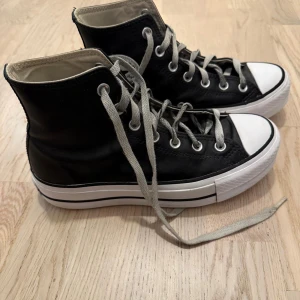 Svarta Converse All Star high tops - Säljer ett par klassiska svarta Converse All Star high tops med vit tå och sula. Skorna har grå snörning, rund tå och är tillverkade i skinn med det ikoniska All Star-märket på sidan och hälen. Perfekt för dig som gillar tidlös streetstil.