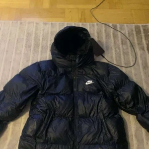 Svart Nike pufferjacka Therma-Fit XL - Svart pufferjacka från Nike i storlek XL barn storlek med Therma-Fit-teknologi. Jackan har hög krage, dragkedja framtill och klassisk Nike-logga på bröstet. Perfekt för kalla dagar och riktigt snygg streetstil. Vinterjacka 