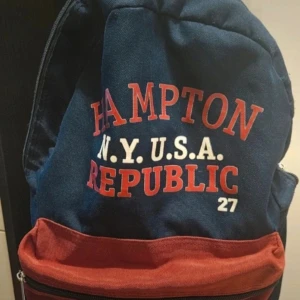 Blå och röd ryggsäck Hampton Republic - Snygg ryggsäck från Hampton Republic i marinblått och rött med tydlig logga framtill. Väskan har ett stort huvudfack, röd frontficka med dragkedja och vadderade axelremmar i rött och blått. Perfekt för skola eller cityhäng.