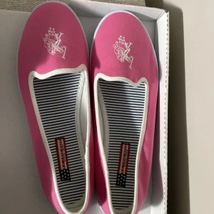 Rosa ballerinaskor från U.S. Polo Assn. - Snygga rosa ballerinaskor från U.S. Polo Assn. med vit kant och vit logga framtill. Skorna har randigt innerfoder i blått och vitt samt platt vit sula. Tillverkade i textilmaterial, perfekta för en chill och färgglad look.