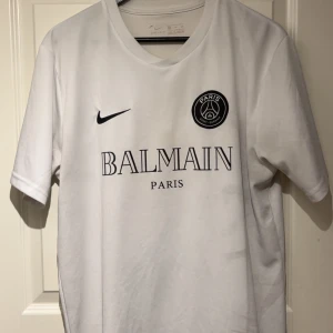 Vit PSG x Balmain fotbollströja Nike - Snygg vit fotbollströja från Nike med Paris Saint-Germain x Balmain tryck på bröstet. Tröjan är kortärmad och tillverkad i Dri-Fit material som andas. Klassisk passform med rund hals och autentisk badge från 2021. Perfekt för dig som gillar exklusiva samarbeten.