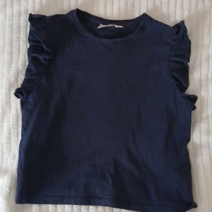 Mörkblå volangtopp från Gina Tricot - Supersöt mörkblå topp från Gina Tricot med volangdetaljer längs axlarna. Toppen är ärmlös och har en klassisk rund halsringning. Tillverkad i mjuk bomull som känns skön mot huden. Perfekt för dig som gillar en enkel men ändå trendig look.💕🩷