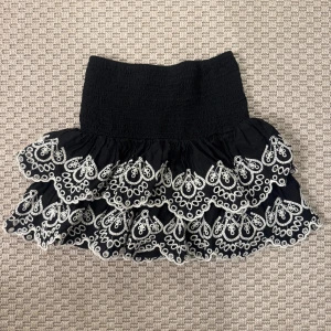 Svart volangkjol med broderi - Supersöt svart kortkjol med bred smock i midjan och dubbla volanger. Kjolen har vita broderade detaljer i ett snyggt mönster längs volangerna som ger en unik look. Perfekt för dig som vill sticka ut lite extra.