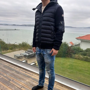 Moncler cardigan  - Svart pufferjacka från Moncler med stickade ärmar och huva. Jackan har dragkedja framtill, quiltad framsida och en ficka med dragkedja på ärmen. Materialet är en mix av ull och polyester, vilket gör den både varm och snygg. Perfekt för kyliga dagar.