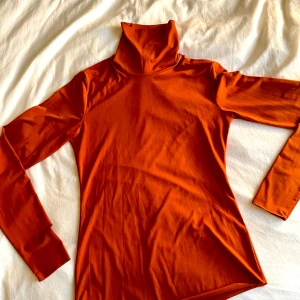 Orange polotröja från Lindex - Snygg orange polotröja från Lindex i stretchigt material. Tröjan har lång ärm och en hög polokrage, perfekt att styla med jeans eller kjol. Tillverkad i mjuk polyesterblandning som sitter skönt på kroppen.