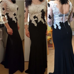 Svart långklänning med spets och blommor - Elegant svart långklänning med figurnära passform, dekorerad med vit och svart spets och blommiga applikationer upptill. Klänningen har transparenta trekvartsärmar och öppen rygg med dragkedja. Perfekt för bal eller festliga tillfällen.