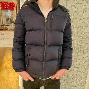Mörkblå dunjacka Polo Ralph Lauren - Snygg mörkblå dunjacka från Polo Ralph Lauren med huva och dragkedja framtill. Jackan har en quiltad look, två sidofickor med dragkedja och en bröstficka. Små defekter på båda ärmarna som knappt syns. Perfekt för kalla dagar och har en klassisk, stilren look. Modellen på bilden är 173 och den är kort i längd och kort i ärmarna