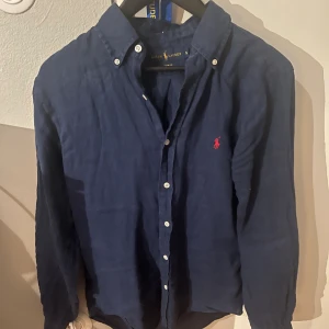 Blå linneskjorta från Ralph Lauren - Snygg marinblå linneskjorta från Ralph Lauren i slim fit. Mycket bra skick, köptes för typ 1 år sen här på plick. Nypris är cirka 2000 kr. Vårt pris 399 kr! Modellen är 183 cm och väger cirka 65 kg. Hör av dig vid minsta fråga!