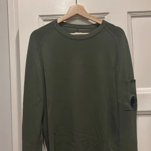 Cp company tröja  - Snygg mörkgrön stickad tröja från C.P. Company med rund hals och lång ärm. Tröjan har en cool detalj med en svart lins på vänster ärm, typiskt för märket. Materialet är mjukt och skönt, perfekt för lager-på-lager under hösten och vintern. Obs den har hål på baksidan!