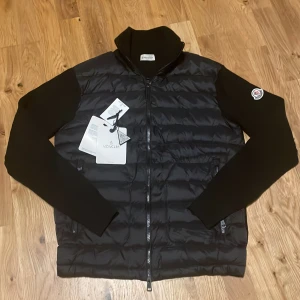 Svart Cardigan från Moncler - Svart Cardigan från Moncler med stickade ärmar och hög krage. Jackan har quiltad front i syntetmaterial, dragkedja och Moncler-logga på vänster ärm. Snygg och stilren design som passar perfekt till höst och vinter.
