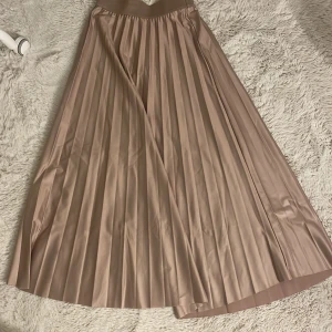 Plisserad beige långkjol - Säljer en snygg plisserad långkjol i beige ton. Kjolen har ett brett midjeband och ett elegant fall med tydliga veck. Perfekt för dig som gillar stilrena och klassiska plagg med lite extra volym. 