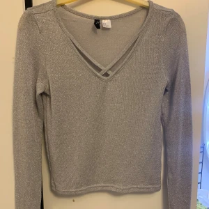 Glittrig silvertröja från H&M Divided - Snygg glittrig silverfärgad tröja från H&M Divided med v-ringning och dekorativa band framtill. Långärmad och i ett stretchigt, mjukt material som känns bekvämt. Perfekt när du vill sticka ut lite extra.