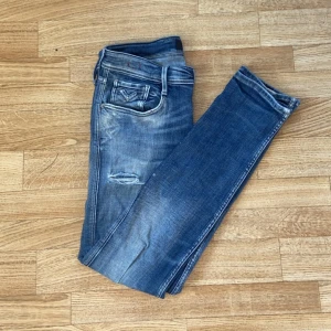 Blå Replay jeans med slitningar - Säljer ett par blå jeans från Replay med snygga slitningar och lätt tvättad look. Modellen har klassisk femficksdesign, normal passform och raka ben. Jeansen är tillverkade i bomull och har detaljerade sömmar vid fickorna.