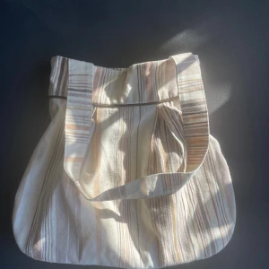 Randig shoppingväska i bomull - En stor och rymlig shoppingväska i bomullstyg med beige, bruna och vita ränder. Väskan har bred axelrem och är helfodrad invändigt. Perfekt för att bära med sig allt man behöver under dagen. Snygg och enkel design som passar till många stilar.