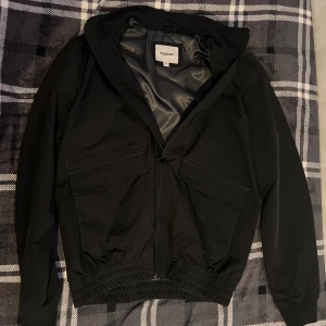 Svart vindjacka från Kappahl med huva - Svart vindjacka från Kappahl med sju fickor i storlek M. Jackan har huva, två stora fickor framtill med lock, dragkedja och elastisk midja. Insidan är fodrad med glansigt tyg. Perfekt för blåsiga dagar och enkel att matcha med olika outfits.