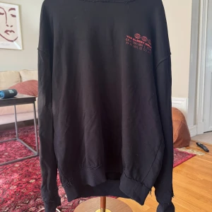 Svart hoodie The Classy Issue - Svart hoodie med coolt rött grafiskt tryck av två ansikten på ryggen och liten röd text på bröstet. Tröjan har huva, ribbade muddar och är långärmad. Materialet känns som mjuk bomull, perfekt för chill dagar. Snygg och unik design som sticker ut.