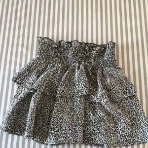 Svartvit blommig volangkjol - Supersöt kort kjol med svartvit blommigt mönster och volanger. Kjolen har en smockad midja som ger en skön och flexibel passform. Perfekt för dig som gillar en feminin och lekfull stil. Materialet känns lätt och luftigt.