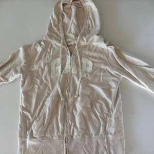 Beige hoodie från GAP med dragkedja - Snygg beige hoodie från GAP med broderad logga framtill. Tröjan har huva med snörning, två stora fickor och dragkedja hela vägen. Tillverkad i mjuk bomull som är skön att ha på sig. Perfekt för chill dagar och enkel att matcha med jeans eller leggings.