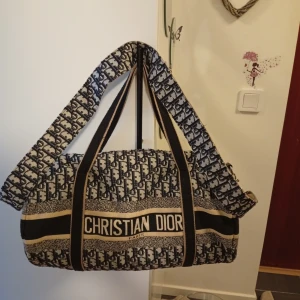 Axelväska med logomönster - Snygg axelväska i tyg med tydligt logomönster i svart och beige. Väskan har bred axelrem med D-tryck, dragkedja upptill och klassisk rundad form. Perfekt för dig som gillar statement-plagg och exklusiv design.