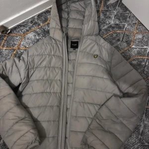 Grå pufferjacka från Lyle & Scott - Säljer en grå pufferjacka från Lyle & Scott med huva och dragkedja. Jackan har det klassiska gula fågel-loggan på bröstet och är quiltad för extra värme. Perfekt för kyliga dagar och har en clean, stilren look.