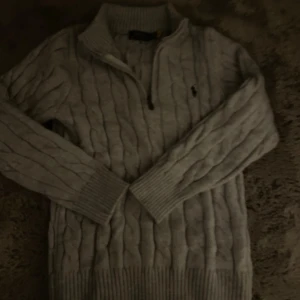 Grå kabelstickad tröja Polo Ralph Lauren - Kabelstickad grå tröja från Polo Ralph Lauren med halv dragkedja och hög krage. Klassisk design med ribbade muddar och diskret broderad logga på bröstet. Perfekt för lager-på-lager och snygg till jeans eller kjol.