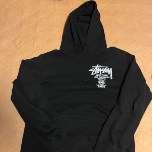Svart Stüssy hoodie med tryck - Svart hoodie från Stüssy i 100% bomull med ikoniskt vitt tryck fram och bak. Klassisk streetwear-look med huva och känguruficka. Perfekt för dig som gillar snygga och bekväma plagg med attityd.