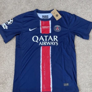 PSG matchtröja Nike blå/röd - Paris Saint-Germain fotbollströja i blått med röd och vit rand längs mitten, officiell logga och Qatar Airways-tryck. Kortärmad modell i lätt funktionsmaterial, perfekt för match eller träning. Champions League-märke på ärmen. frakatar inom 24 timmar.