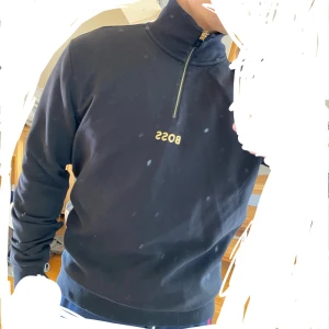 Svart BOSS half zip sweatshirt - Helt ny Svart sweatshirt från Hugo Boss (köpt från Zalando) med guldfärgad dragkedja och broderad BOSS-logga på bröstet. Tröjan har half zip, hög krage och ribbade muddar. Perfekt för en clean och stilren look. 