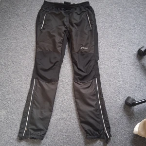 Svarta längdskidbyxor från Skigo, strl M - Svarta längdskidbyxor från Skigo med elastisk midja och dragkedjor längs benen. Byxorna har förstärkta partier på knän och vader, samt praktiska fickor med dragkedja. Perfekta för träning utomhus under kyliga dagar.