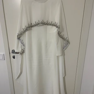 Vit långklänning med silverdetaljer - Vit långklänning med raka långa ärmar och exklusiva silverfärgade paljettdetaljer längs överdelen och ärmarna. Klänningen har en elegant och flytande siluett, tillverkad i ett lätt material som ger ett lyxigt intryck. Perfekt för dig som vill sticka ut med glittrande detaljer.